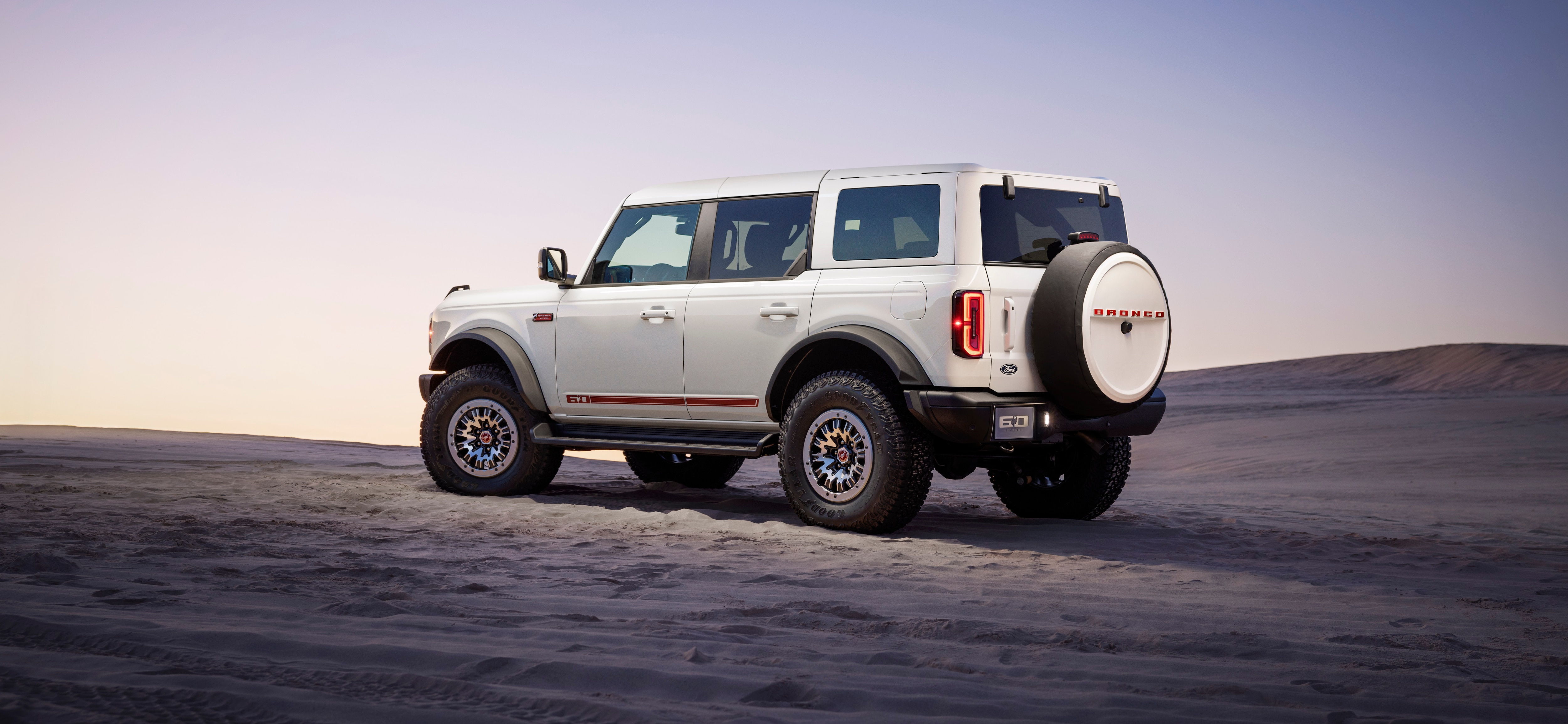 2026 Ford Bronco 60th Anniversary Package side view showing a white vehicle with red accents. Vehicle is on the sand with a purple and yellow dusk sky overhead.